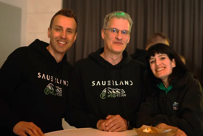 Drei lächelnde Menschen mit Sauerland- und P-Weg-Team-Logos auf der Kleidung stehen eng zusammen. Im Hintergrund sind weitere Personen an Tischen zu sehen.