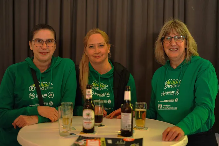 Drei lächelnde Frauen mit grünen T-Shirts und Logos von Sponsoren stehen nebeneinander und halten Bierflaschen. Im Hintergrund ein grauer Vorhang.