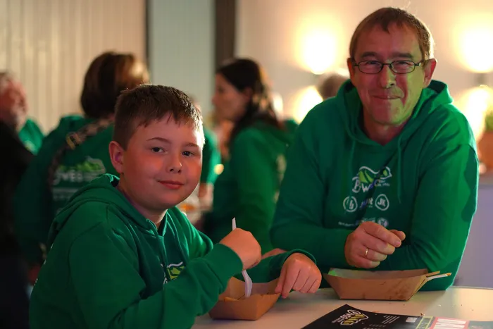 Ein Junge und ein Mann in grünen Hoodies sitzen an einem Tisch und essen Pommes aus einer Papiertüte. Im Hintergrund sind weitere Personen in grünen T-Shirts zu sehen.
