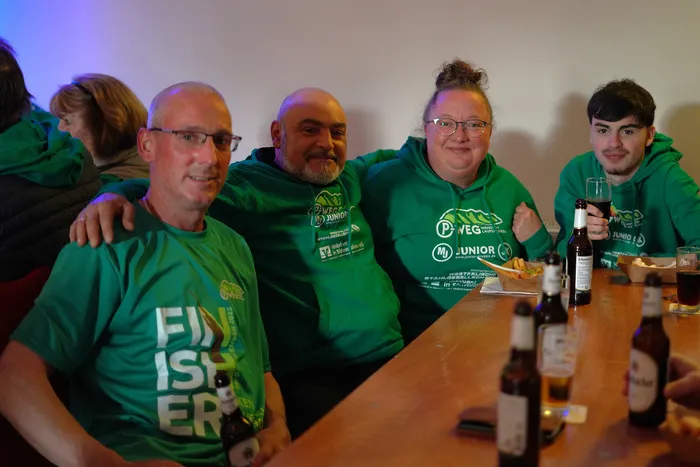 Fünf Personen in grünen T-Shirts sitzen an einem Holztisch mit Getränken und Snacks, während sie gemeinsam in die Kamera lächeln. Im Hintergrund ist ein Teil einer Person zu sehen, die über die Schulter schaut.