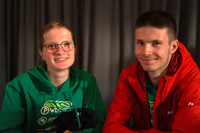 Zwei junge Erwachsene lächeln in die Kamera. Die Frau links trägt eine grüne Jacke mit Logos, der Mann rechts eine rote Jacke.