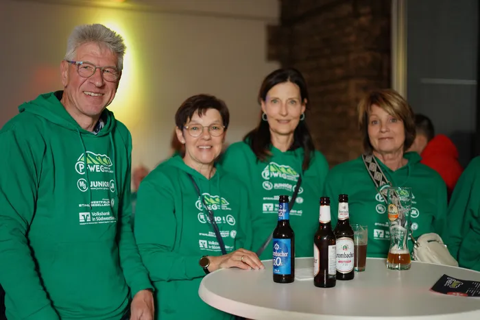 Fünf lächelnde Menschen in grünen T-Shirts stehen um einen runden Tisch mit Bierflaschen und Gläsern. Sie tragen Kleidung mit dem Logo des P-Wegs und Sponsoren.