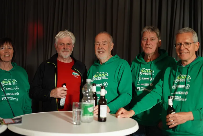 Sechs ehrenamtliche Helfer des P-Wegs stehen lächelnd vor einer dunklen Wand und einem Tisch mit Getränken. Sie tragen grüne Pullover mit dem P-Weg-Logo und den Namen der Sponsoren.