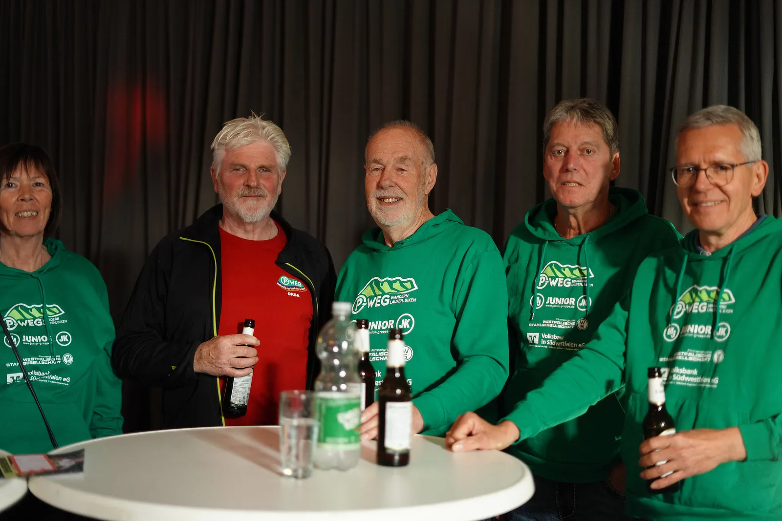 Sechs ehrenamtliche Helfer des P-Wegs stehen lächelnd vor einer dunklen Wand und einem Tisch mit Getränken. Sie tragen grüne Pullover mit dem P-Weg-Logo und den Namen der Sponsoren.