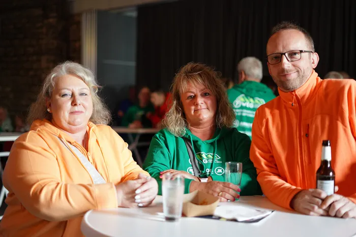Drei lächelnde Menschen sitzen an einem Tisch, eine Frau in Orange und eine in Grün, ein Mann in Orange mit Brille. Im Hintergrund weitere Personen.