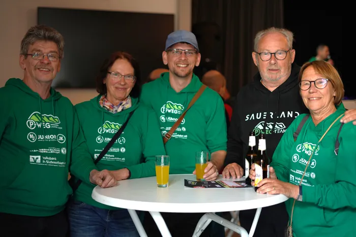 Fünf lächelnde Menschen sitzen an einem runden Tisch mit Getränken, alle tragen grüne Kapuzenpullover mit dem Aufdruck „P-Weg“ und verschiedenen Logos.