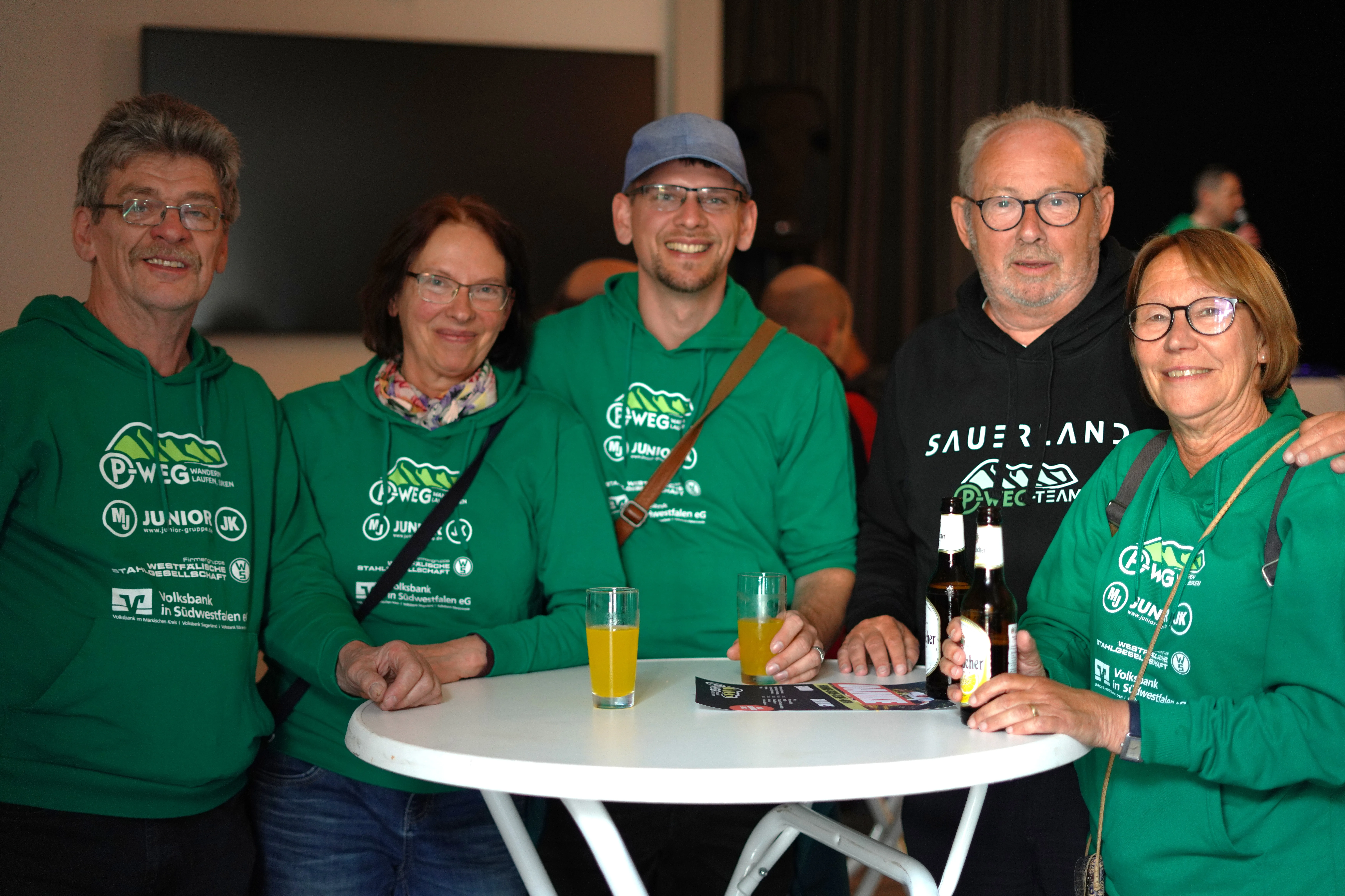 Fünf lächelnde Menschen sitzen an einem runden Tisch mit Getränken, alle tragen grüne Kapuzenpullover mit dem Aufdruck „P-Weg“ und verschiedenen Logos.