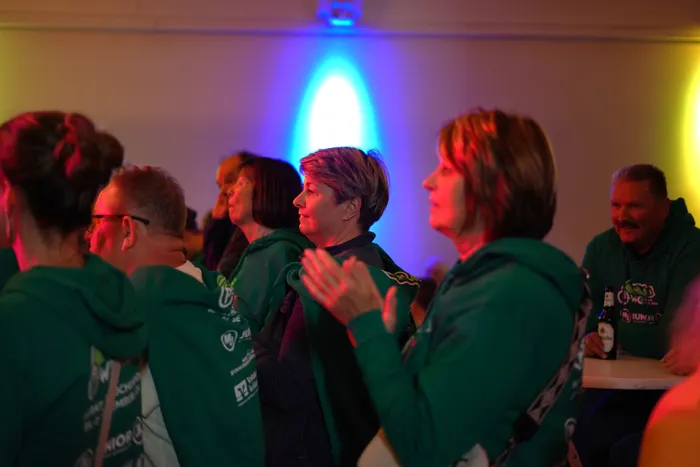 Mehrere Personen in grünen Hoodies sitzen oder stehen in einem Raum mit farbigem Licht und applaudieren.