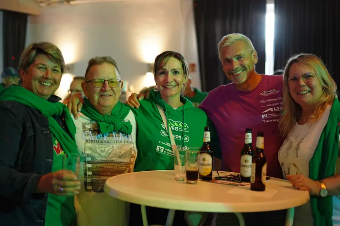 Fünf lächelnde Menschen stehen eng zusammen vor einem Tisch mit Bierflaschen. Sie tragen grüne Kleidung und Accessoires, einige halten Flaggen. Im Hintergrund sind weitere Personen und eine gemütliche Raumgestaltung zu sehen.
