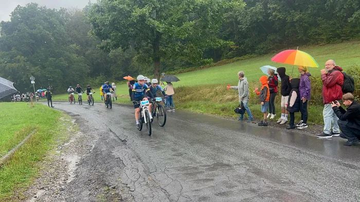Radfahrer bei einem Rennen auf einer nassen Straße, gesäumt von Zuschauern mit bunten Regenschirmen in einer bewaldeten Landschaft.