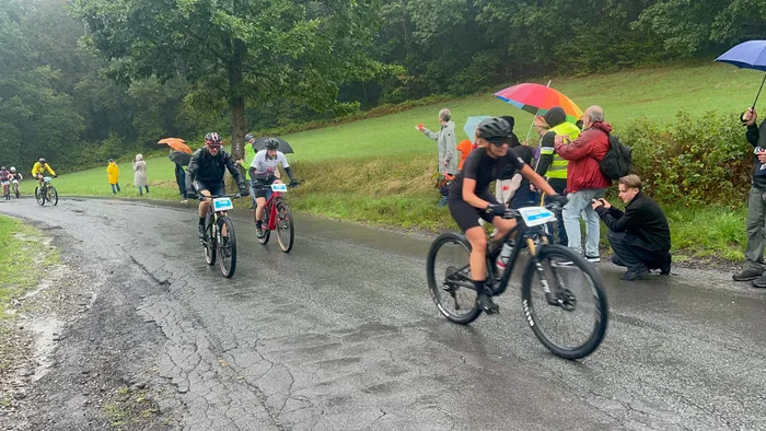 Mountainbiker fahren auf einer nassen Straße an Zuschauern mit bunten Regenschirmen vorbei.