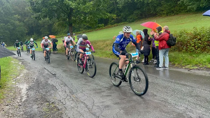 Radfahrer fahren auf einer nassen Straße durch ein Waldgebiet, während Zuschauer am Rand mit Regenschirmen zusehen.