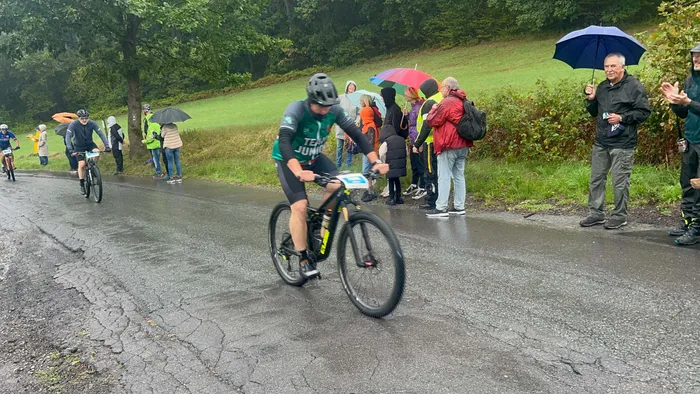 Ein Radfahrer in grünem Trikot fährt auf einer nassen Asphaltstraße an Zuschauern mit Regenschirmen vorbei.