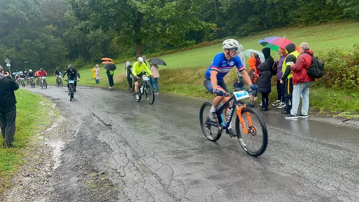 Ein Radfahrer in blauem Trikot fährt auf einer nassen Straße an Zuschauern mit Regenschirmen vorbei.