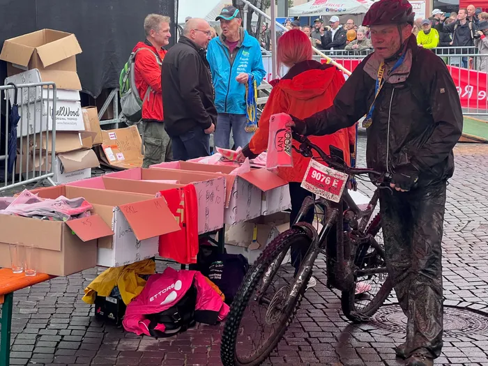 Ein schlammiger Mountainbiker mit rotem Helm steht neben seinem Fahrrad an einem Tisch mit Kartons voller pinkfarbener Kleidung.