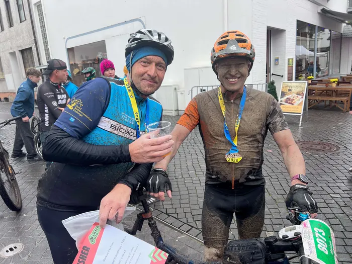 Zwei schlammverschmierte Radfahrer mit Medaillen vom P-Weg-Sonntag 2026 lächeln neben ihren Fahrrädern.