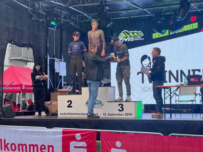 Siegerehrung beim P-Weg-Marathonwochenende 2026 auf einer Bühne, bei der ein Mann Gewinnern auf einem Podium eine Auszeichnung überreicht.
