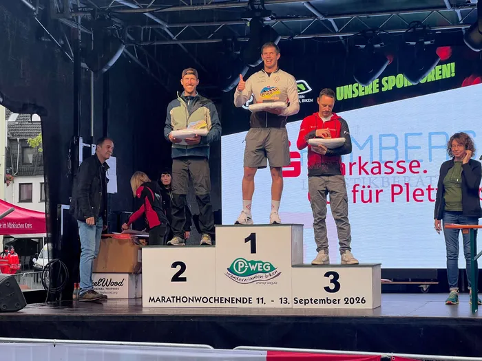 Drei Männer stehen auf einem Siegerpodium beim P-Weg-Marathonwochenende 2026 und halten Teller in den Händen.