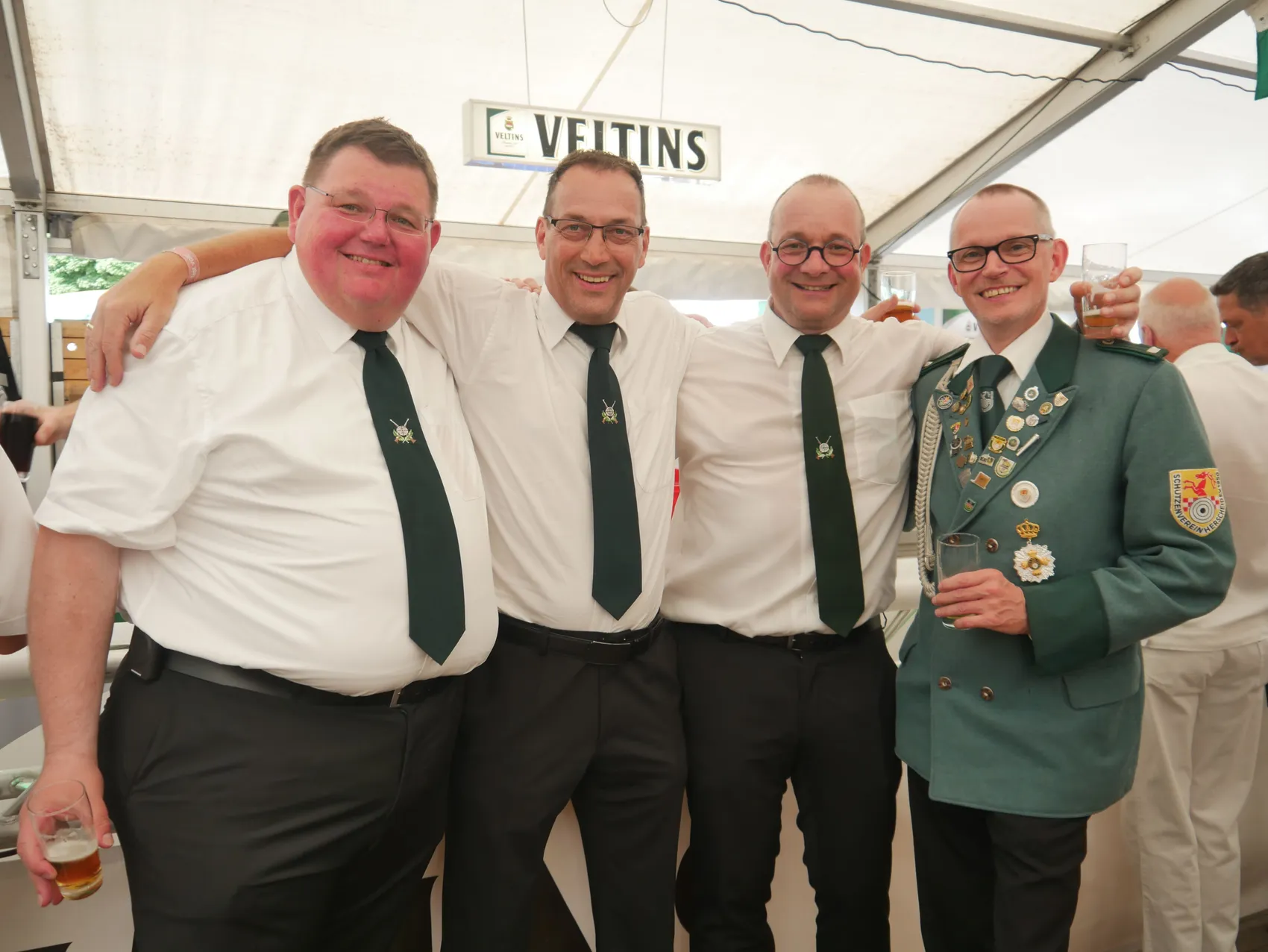Vier lächelnde Männer beim Schützenfest, drei in weißen Hemden mit grünen Krawatten und einer in einer grünen Uniform mit Orden.