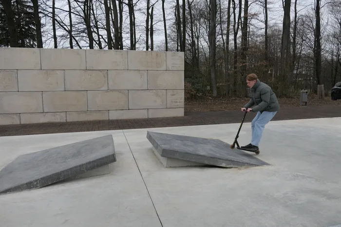 Ein Jugendlicher fährt mit einem Tretroller über eine Rampe in einem Skatepark, im Hintergrund eine Mauer und Bäume.