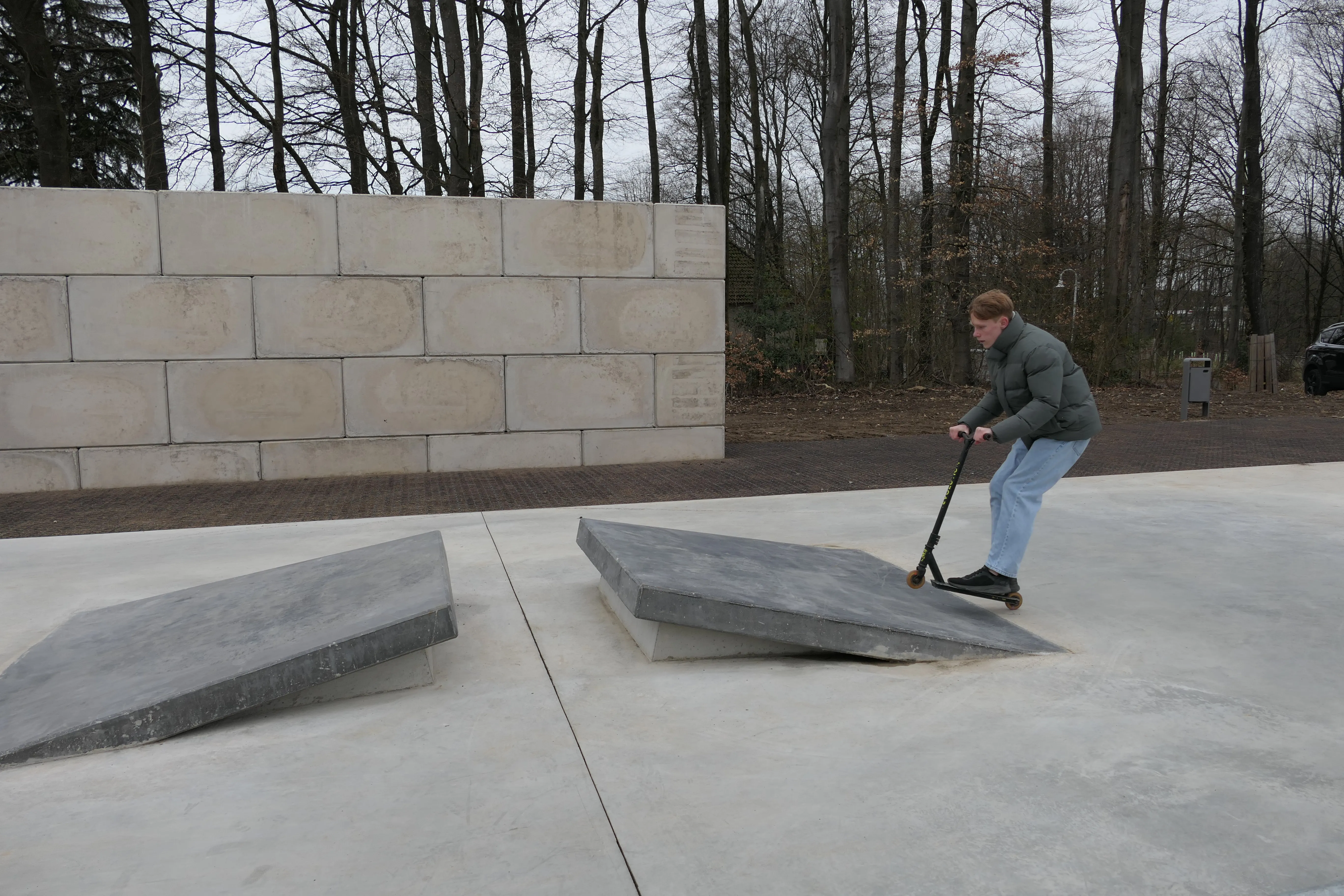 Ein Jugendlicher fährt mit einem Tretroller über eine Rampe in einem Skatepark, im Hintergrund eine Mauer und Bäume.