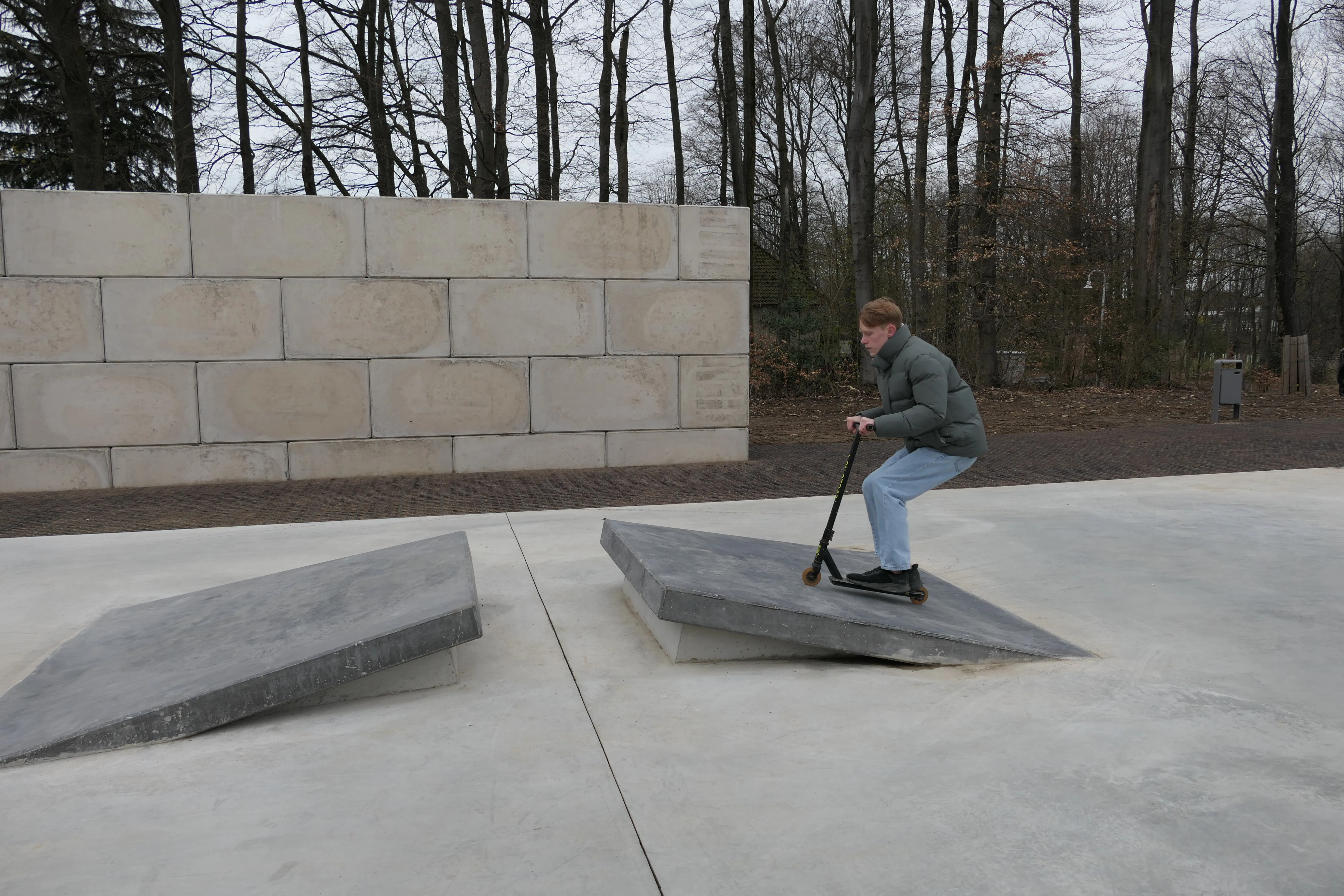Ein Jugendlicher fährt mit einem Tretroller über eine Rampe in einem Park. Im Hintergrund eine Mauer und kahle Bäume.