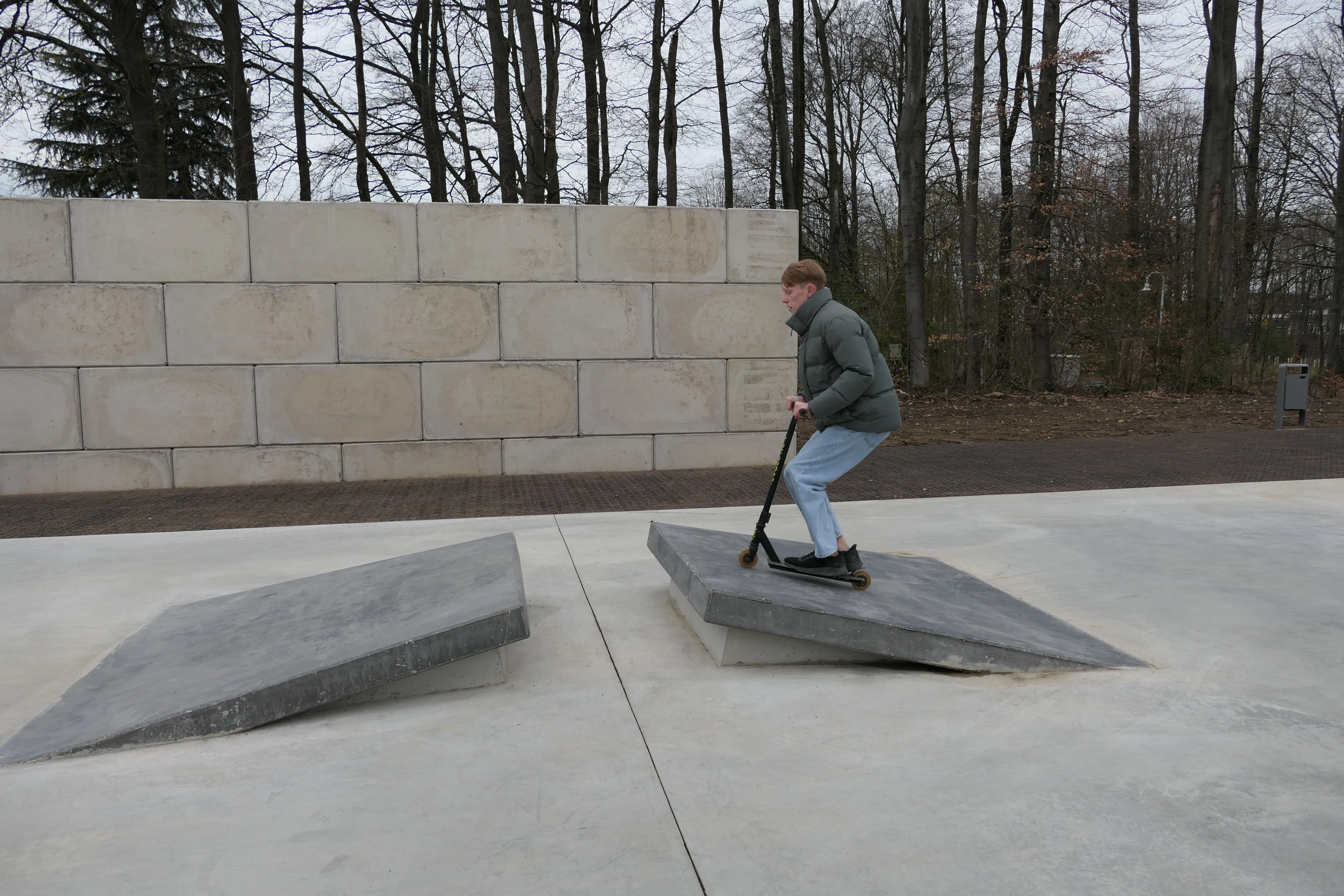 Ein Jugendlicher fährt mit einem Tretroller über eine Rampe in einem Skatepark, im Hintergrund eine Mauer und kahle Bäume.
