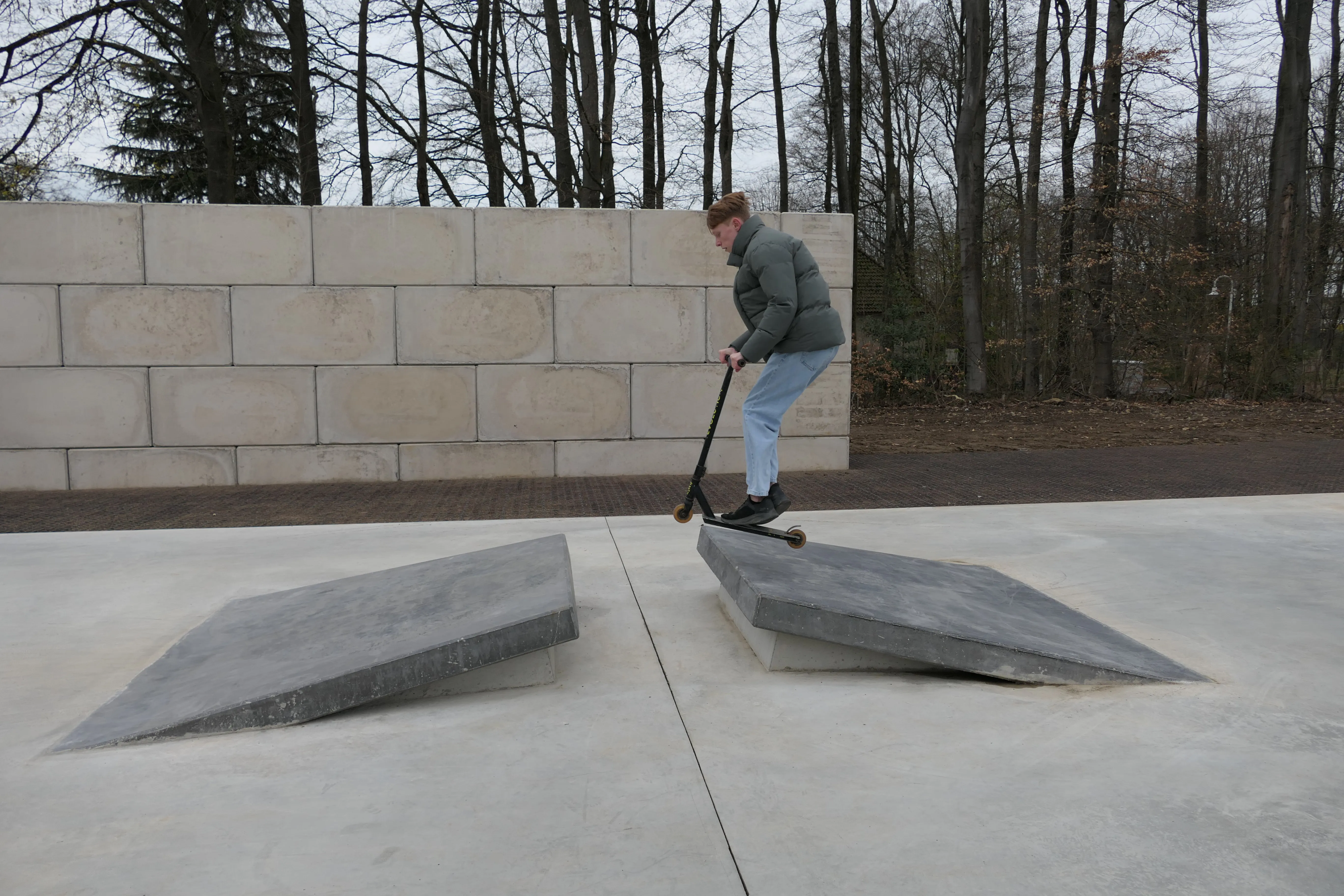 Ein junger Mann fährt mit einem Tretroller über eine schräge Betonfläche in einem Park. Im Hintergrund eine Mauer und kahle Bäume.