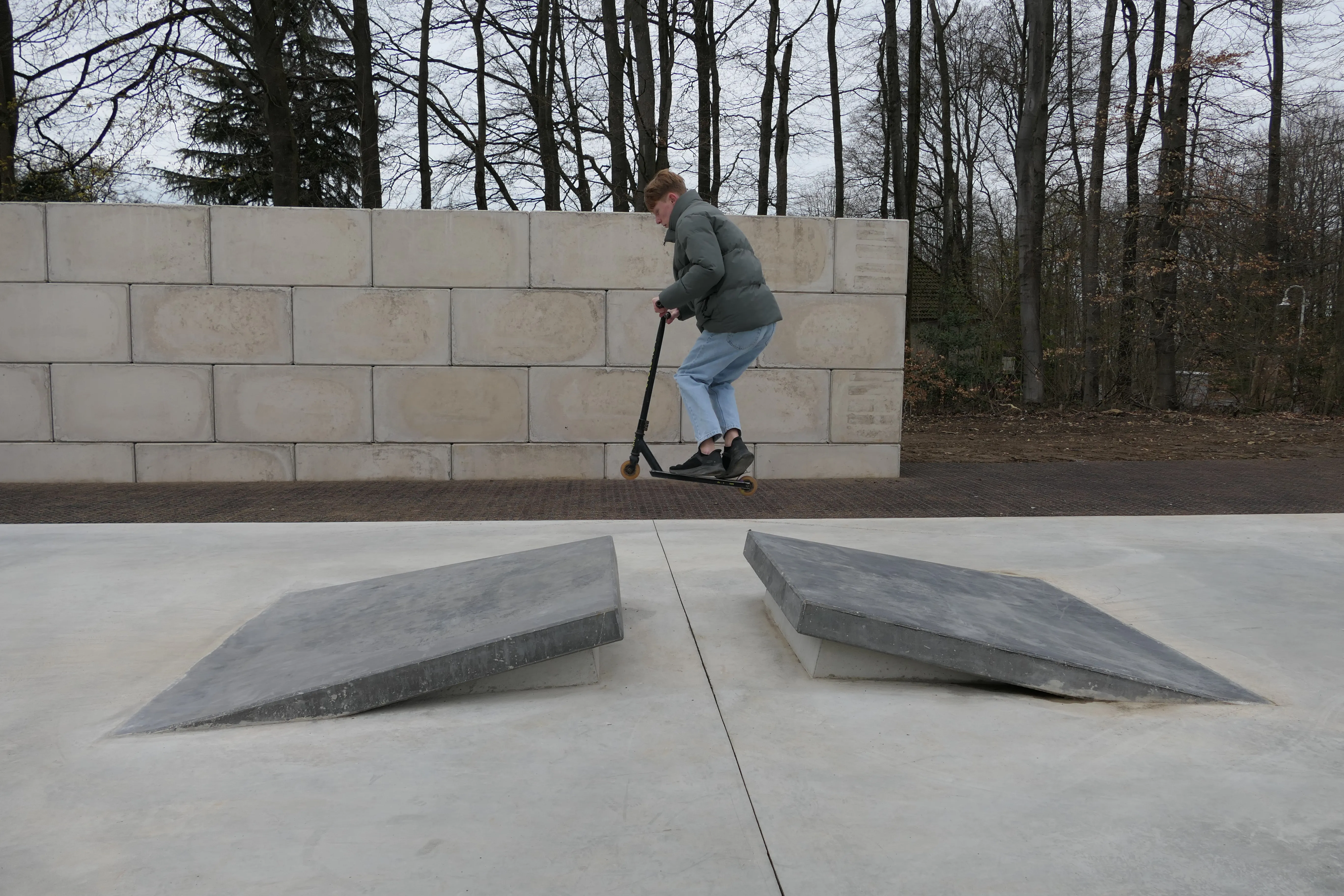 Ein junger Mann fährt mit einem Tretroller über zwei schräge Betonplatten in einem Park.