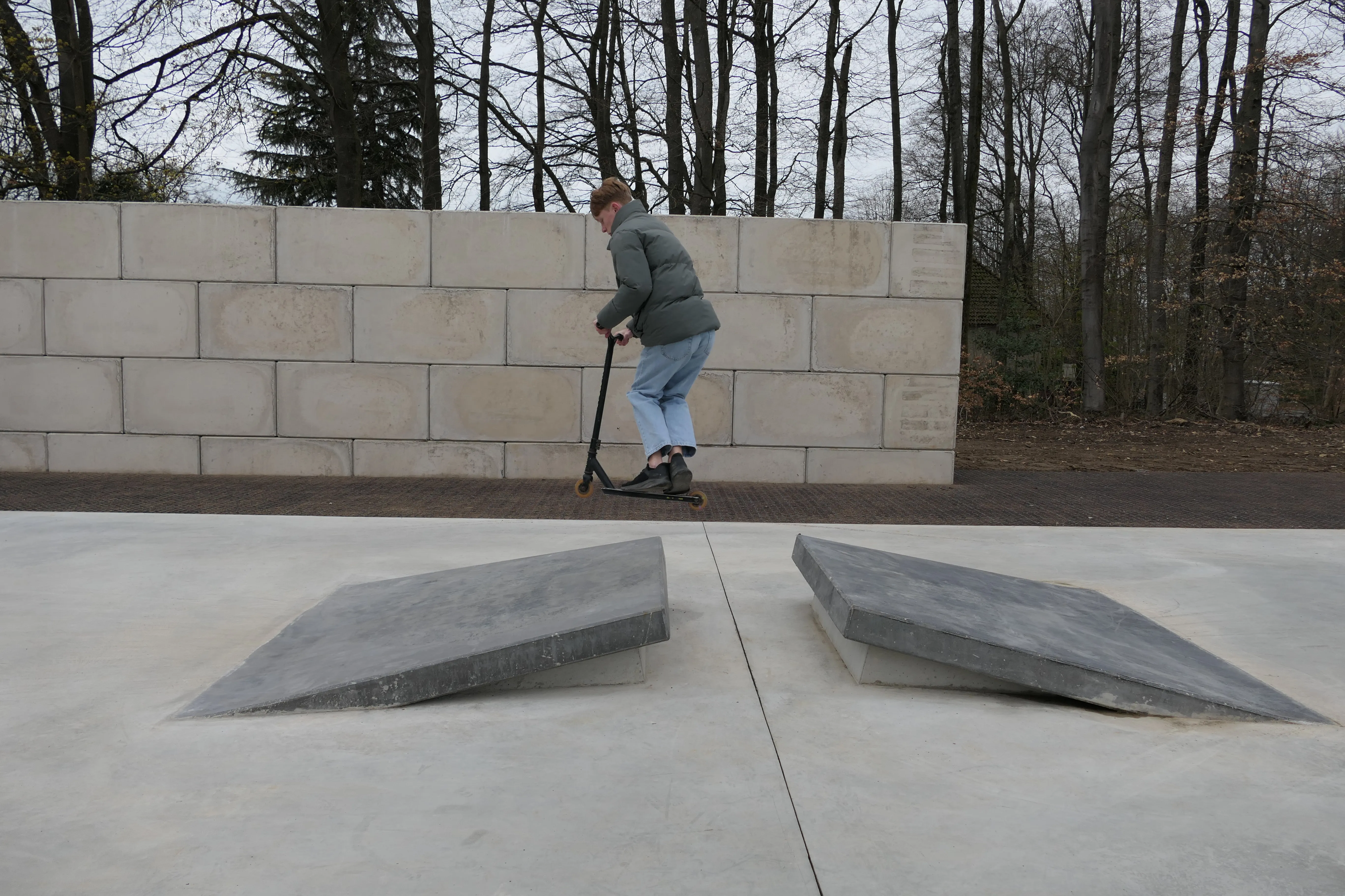 Ein junger Mann fährt mit einem Roller über eine schräge Betonfläche in einem Park.