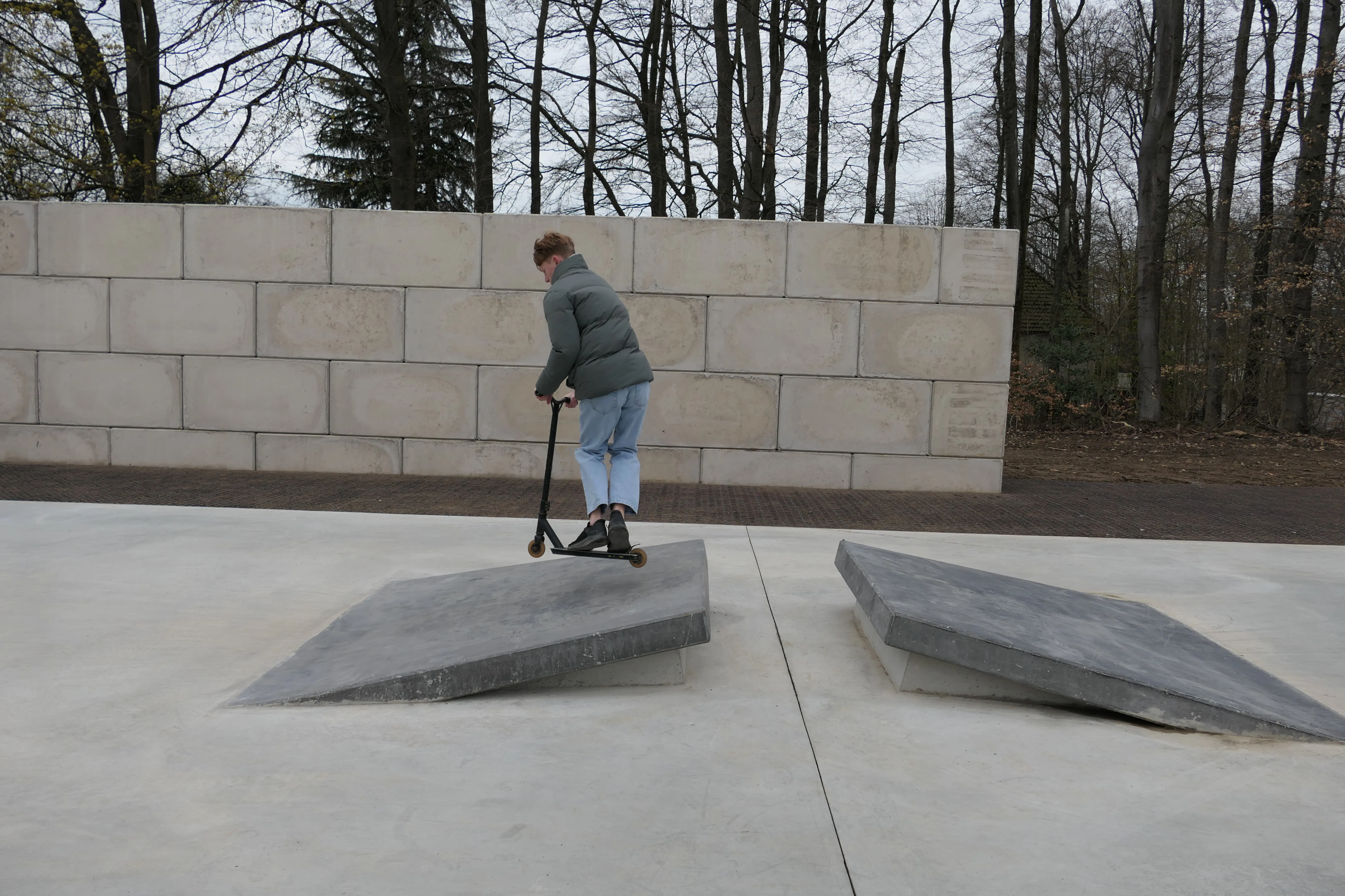 Ein Jugendlicher fährt mit einem Tretroller über eine Betonschanze in einem Skatepark, Bäume im Hintergrund.