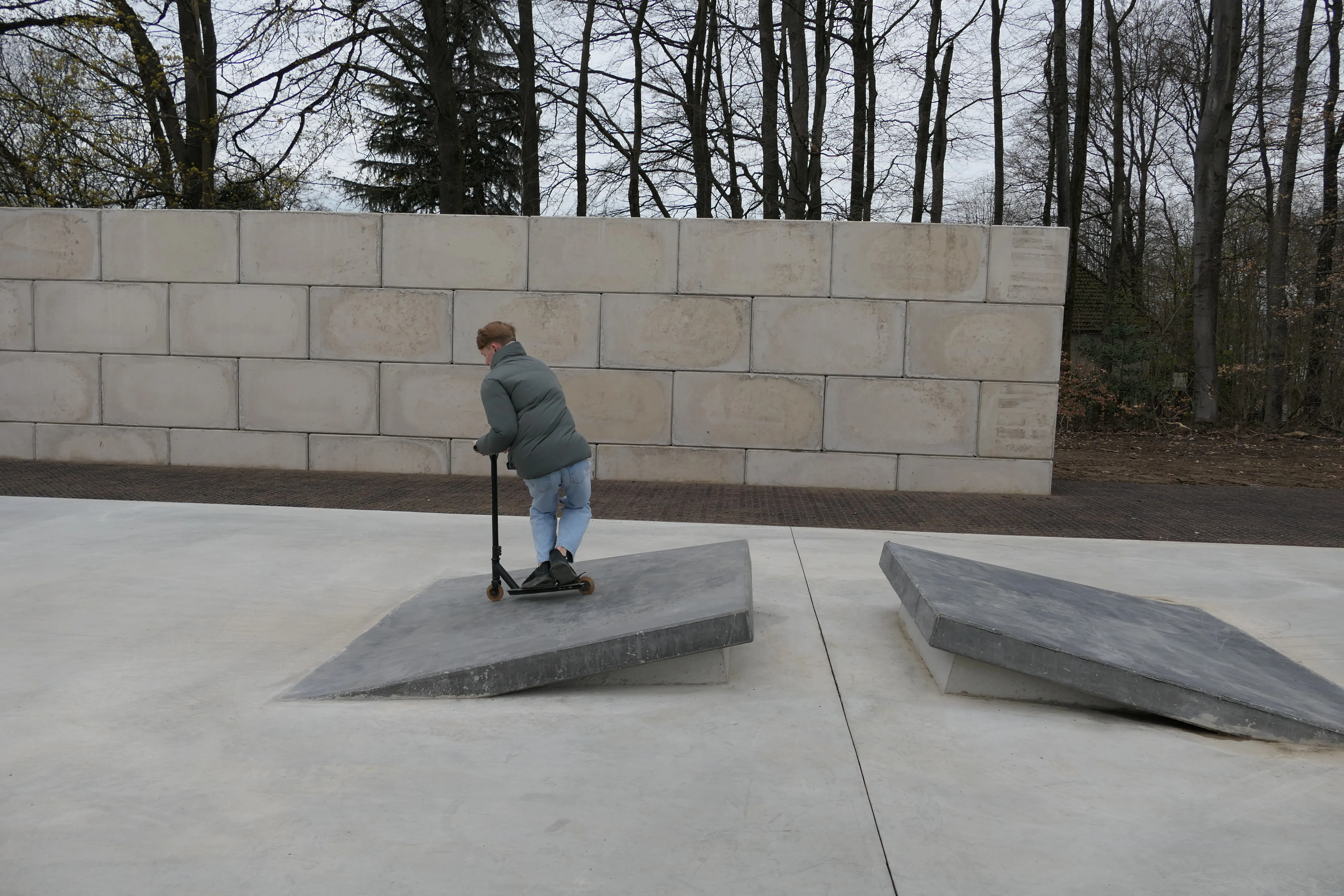 Eine Person fährt mit einem Tretroller über eine schräge Betonfläche in einem Park. Im Hintergrund eine Mauer und Bäume.