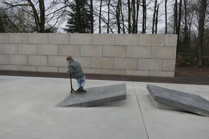 Eine Person fährt mit einem Roller über eine schräge Betonfläche in einem Park. Im Hintergrund eine hohe Mauer und Bäume.
