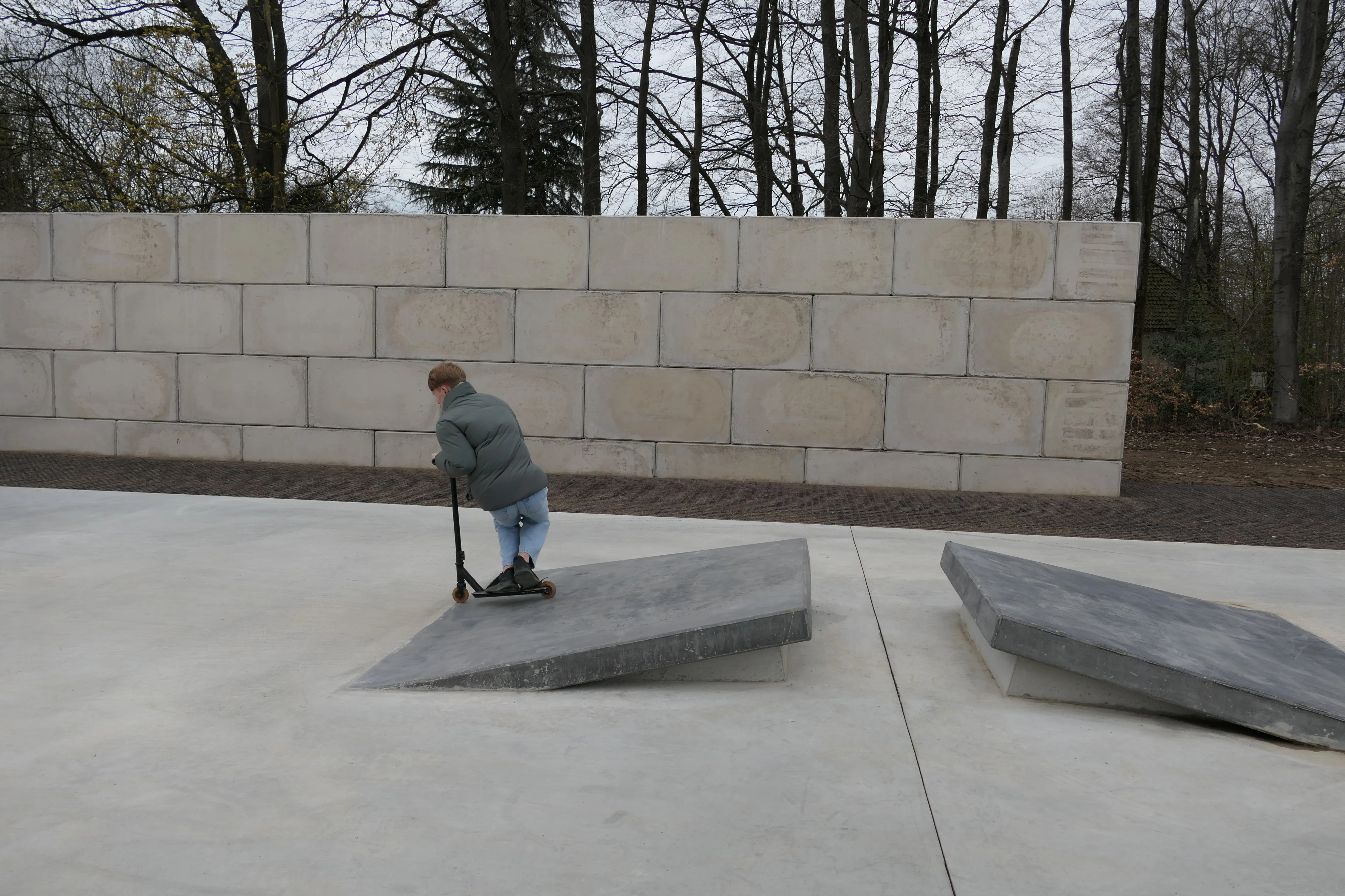 Eine Person fährt mit einem Roller über eine schräge Betonfläche in einem Park. Im Hintergrund eine hohe Mauer und Bäume.