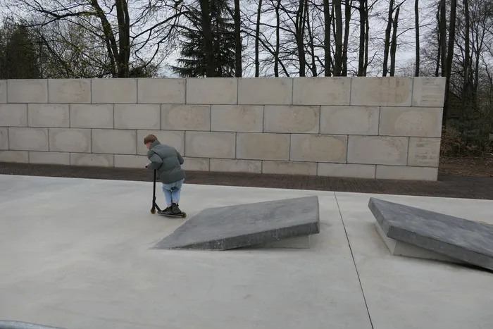 Ein Jugendlicher fährt mit einem Tretroller über eine schräge Betonfläche in einem Park. Im Hintergrund eine hohe Mauer und Bäume.