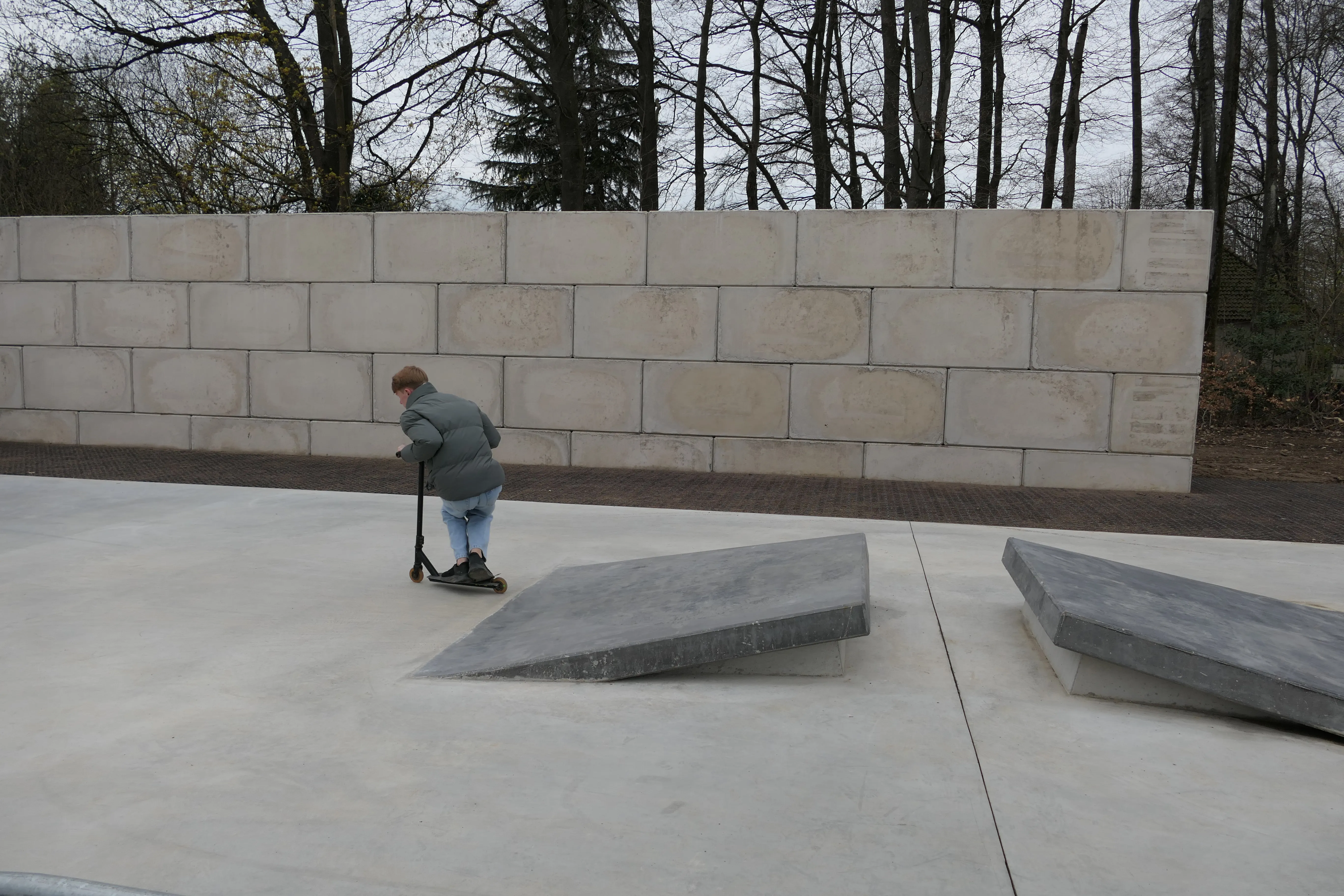 Ein Jugendlicher fährt mit einem Tretroller über eine schräge Betonfläche in einem Park. Im Hintergrund eine hohe Mauer und Bäume.