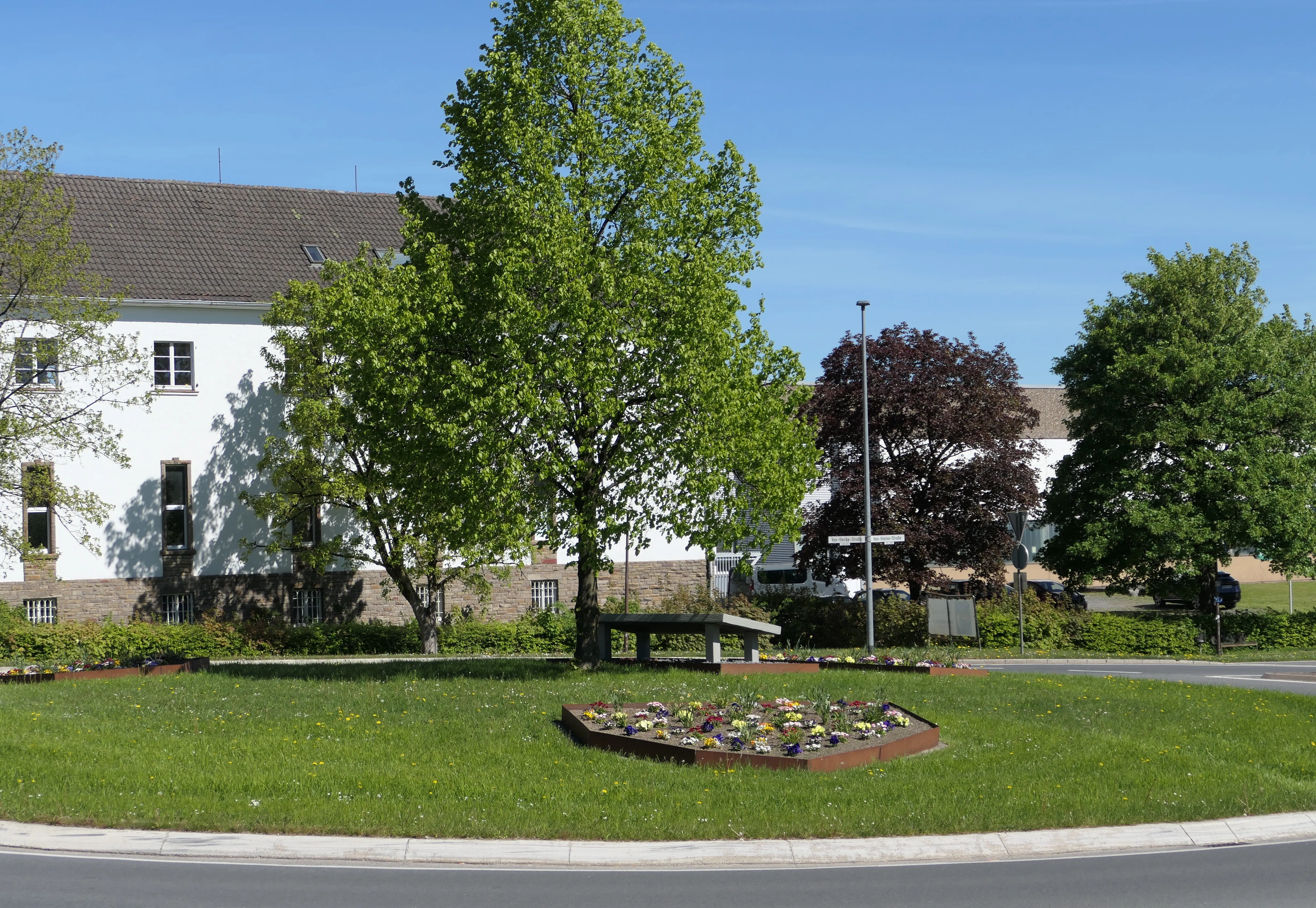 Kreisverkehr mit bunt bepflanztem Blumenbeet im Vordergrund, dahinter Gebäude und Bäume unter blauem Himmel.