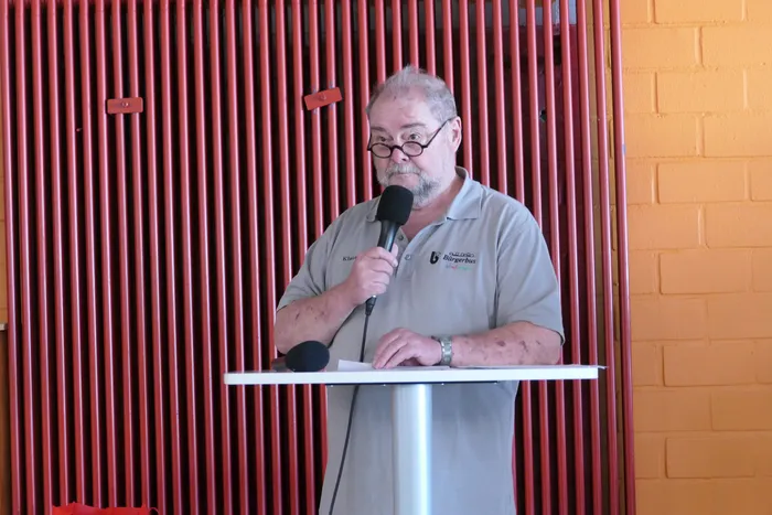Ein Mann mit Brille und khaki-farbenem Polohemd spricht in ein Mikrofon an einem Rednerpult. Im Hintergrund eine rote Metallgitterwand und eine Backsteinmauer.