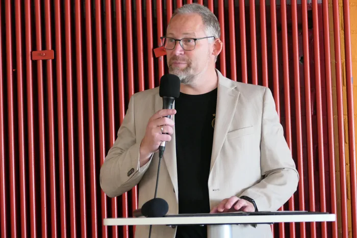 Ein Mann mit Brille und beigem Sakko spricht in ein Mikrofon an einem Pult. Im Hintergrund eine rote Lamellenwand.
