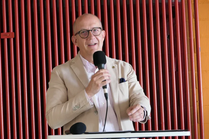 Ein Mann mit Glatze und Brille hält ein Mikrofon und spricht. Er trägt einen beigen Blazer und ein weißes Hemd. Im Hintergrund ist ein rotes Gitter zu sehen.