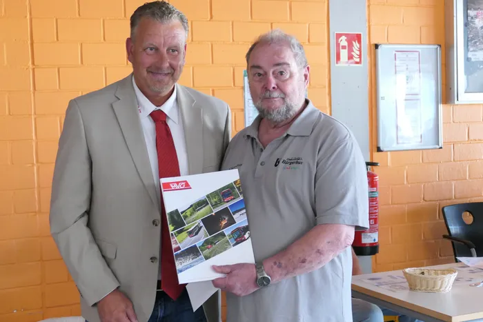 Zwei Männer stehen nebeneinander und halten ein Fotoalbum. Der Mann links trägt einen Anzug mit roter Krawatte, der rechte ein grünes Poloshirt mit Aufschrift 