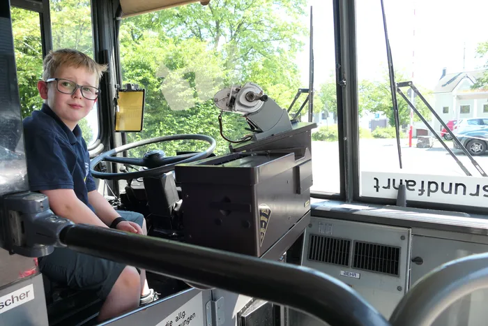 Junge mit Brille sitzt am Steuer eines Oldtimerbusses. Im Hintergrund ist durch die Fenster die Straße zu sehen.