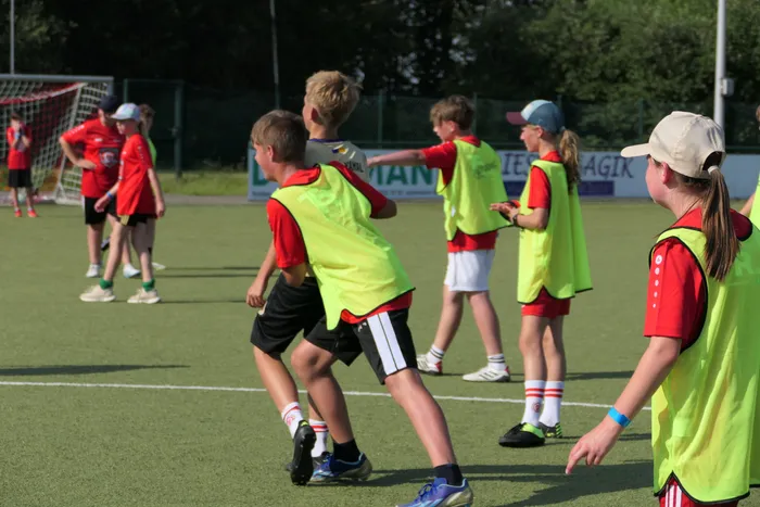 Kinder trainieren Fußball auf einem Kunstrasenplatz. Einige tragen gelbe Trainingswesten, andere rote Trikots. Im Hintergrund ist ein Fußballtor und Werbebanden zu sehen.