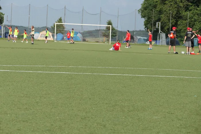 Fußballcamp des TuS Ennepe mit Kindern und Trainern auf einem grünen Kunstrasenplatz. Einige Spieler laufen dem Ball hinterher, andere stehen oder sitzen am Spielfeldrand. Im Hintergrund ein Fußballtor und Bäume.