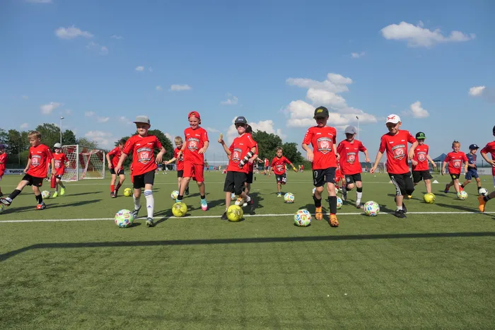 Eine Gruppe Jugendlicher in roten Trikots trainiert auf einem grünen Fußballfeld. Sie üben Dribbling mit mehreren Fußbällen.