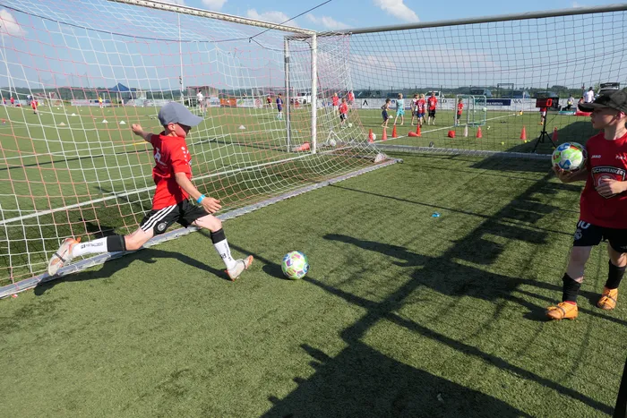 Ein Junge schießt während eines Fußballcamps auf das Tor, während ein anderer Junge mit rotem Trikot und Handschuhen bereit steht. Im Hintergrund sind weitere Kinder und Trainingsutensilien auf einem Fußballfeld zu sehen.