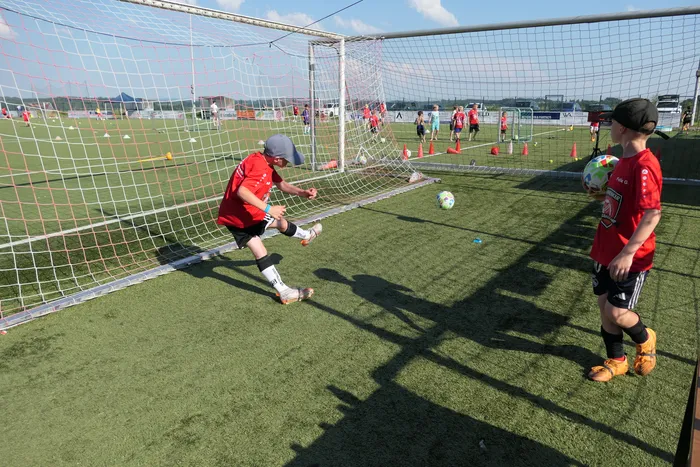 Ein Jugendlicher schießt während eines Fußballcamps auf ein kleines Tor. Ein zweiter Jugendlicher steht bereit, den Ball zu verteidigen. Im Hintergrund trainieren weitere Kinder auf einem Fußballfeld.