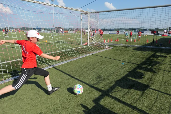 Ein Jugendlicher in rotem Trikot und schwarzer Hose schießt auf einem Fußballfeld gegen einen Ball. Im Hintergrund sind weitere Spieler und Trainingshütchen zu sehen.