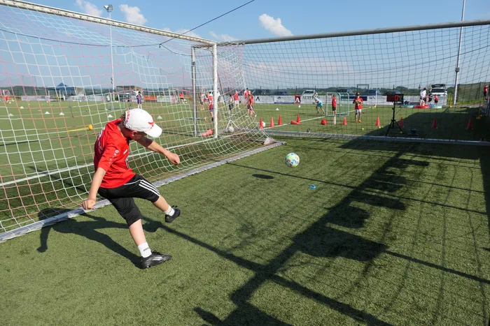 Ein Jugendlicher in rotem Trikot und schwarzen Shorts schießt auf ein Fußballtor auf einem grünen Kunstrasenplatz. Im Hintergrund sind weitere Spieler und Trainingsutensilien zu sehen.