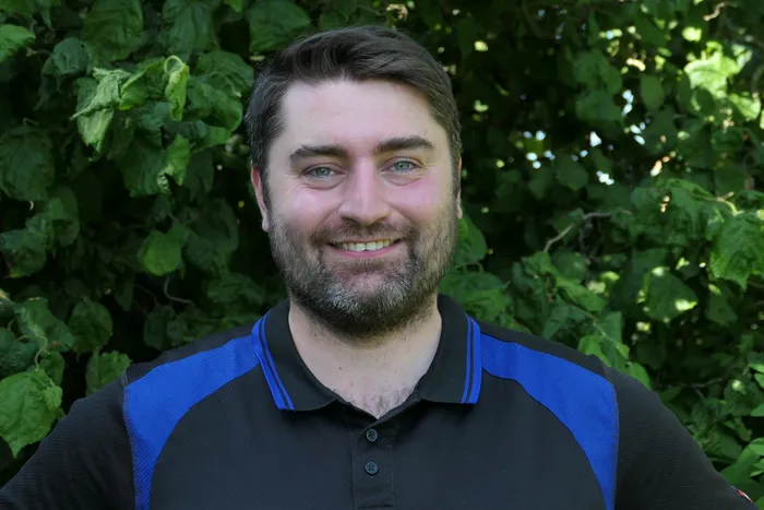 Ein lächelnder Mann mit dunklem Haar und Bart trägt ein blau-schwarzes Poloshirt vor einem grünen Blätterhintergrund. Er ist Yannick Prümers, der neue Leiter des Forstbetriebes Halver.